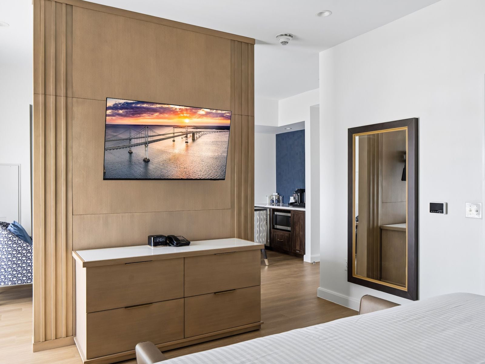 Une chambre moderne avec un grand lit, une commode en bois, un tableau représentant un coucher de soleil et une vue sur la cuisine.