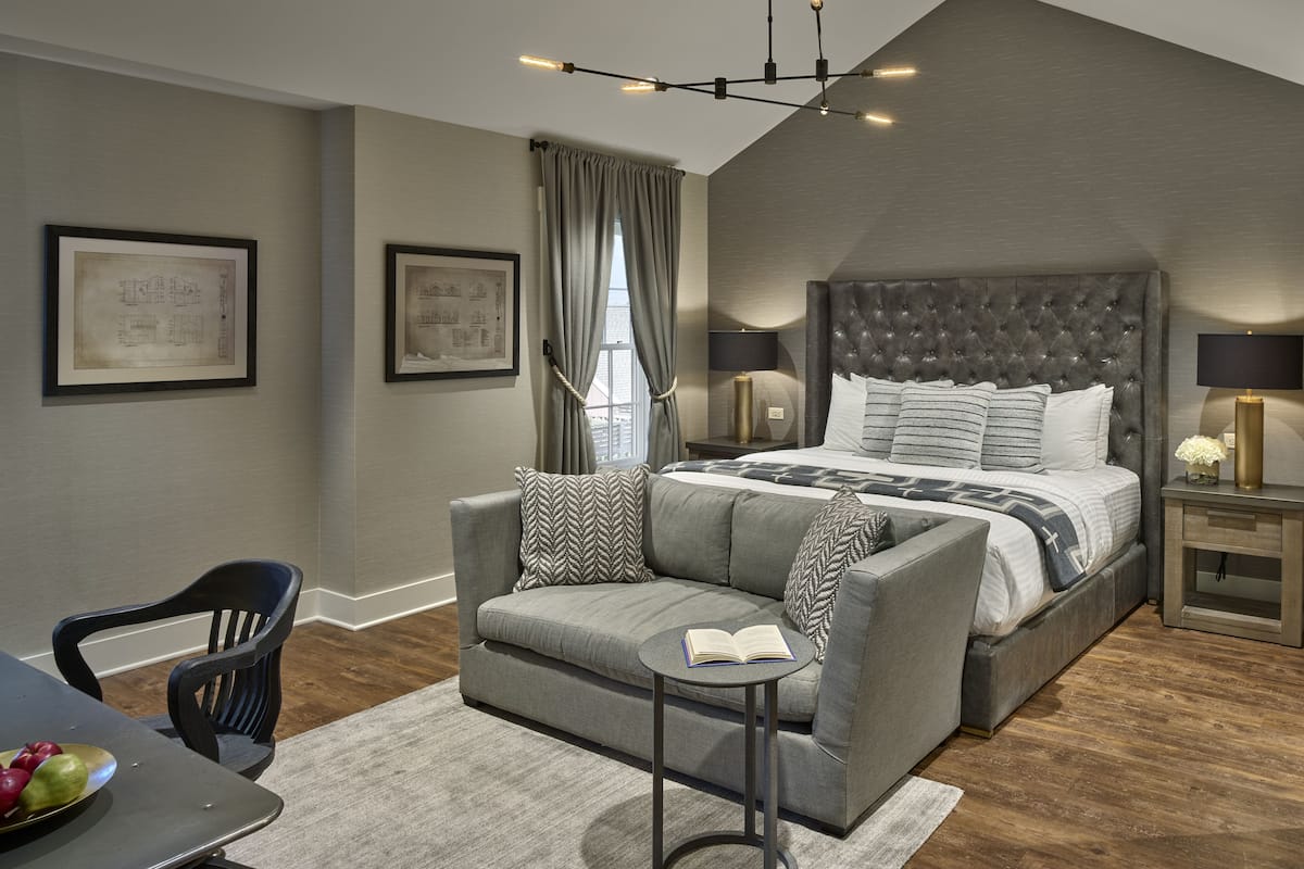 A modern bedroom with a large bed, a grey sofa, a small table with a book, and a dining area with a chair and a bowl of fruit.