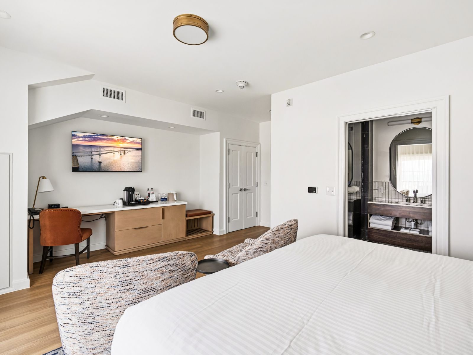 A modern hotel room with a large bed, a desk with a computer, a chair, and a view of the sea through the window.