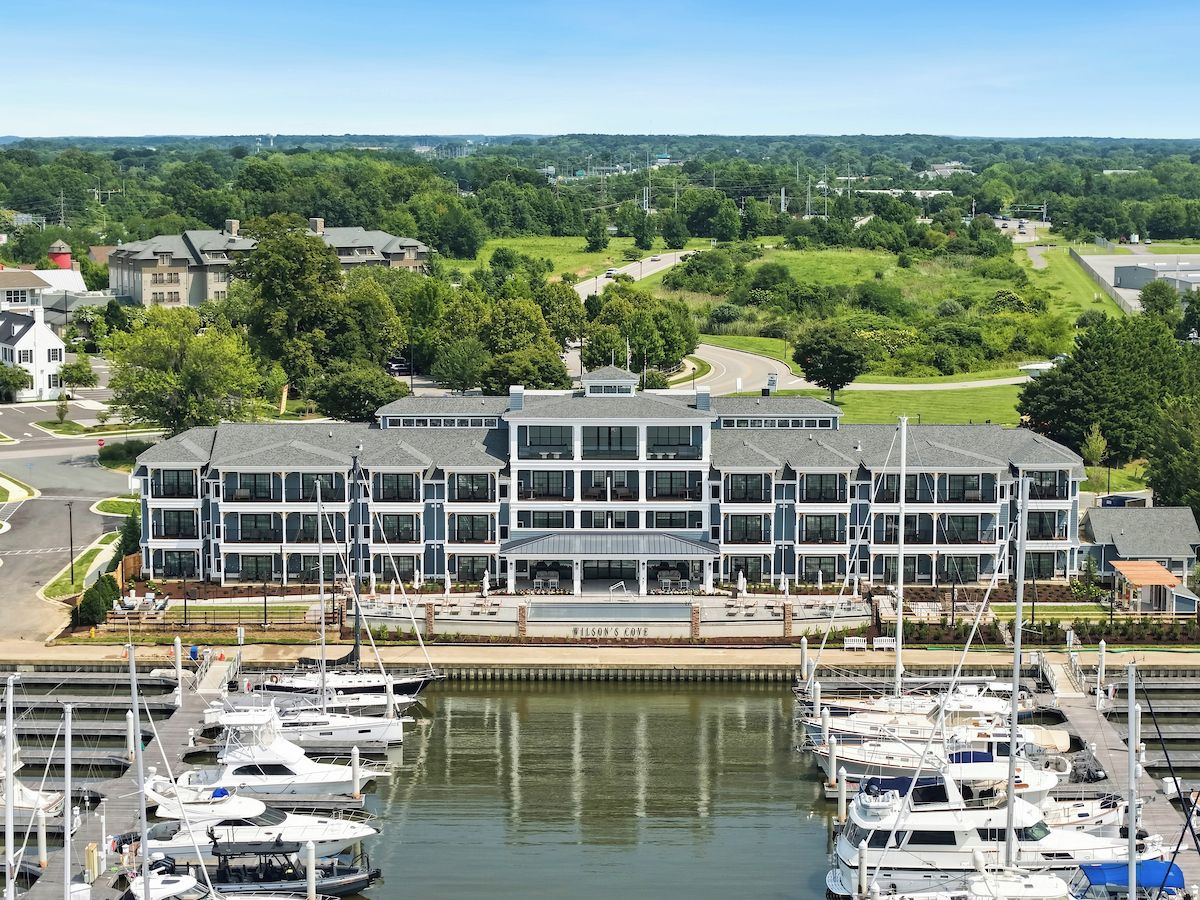 A marina with multiple boats docked, a large building with a sign reading 'Wilson's Cove,' and a surrounding landscape with greenery and other buildings.