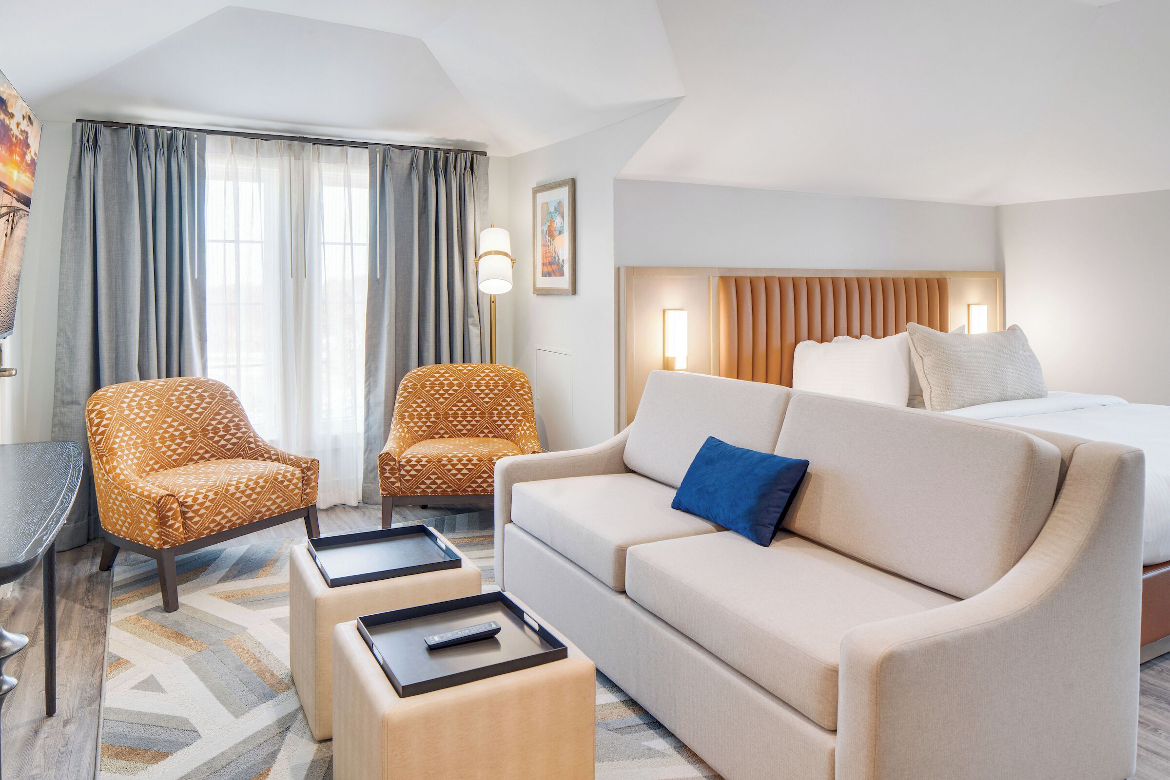 Ein Hotelzimmer mit einer Couch, einem Sessel und einem Bett mit Kopfteil. Es gibt einen Schreibtisch mit einem Stuhl und einer Lampe.