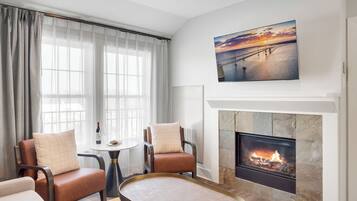 A modern living room with a fireplace, a large window with sheer curtains, and a painting of a sunset over a bridge.