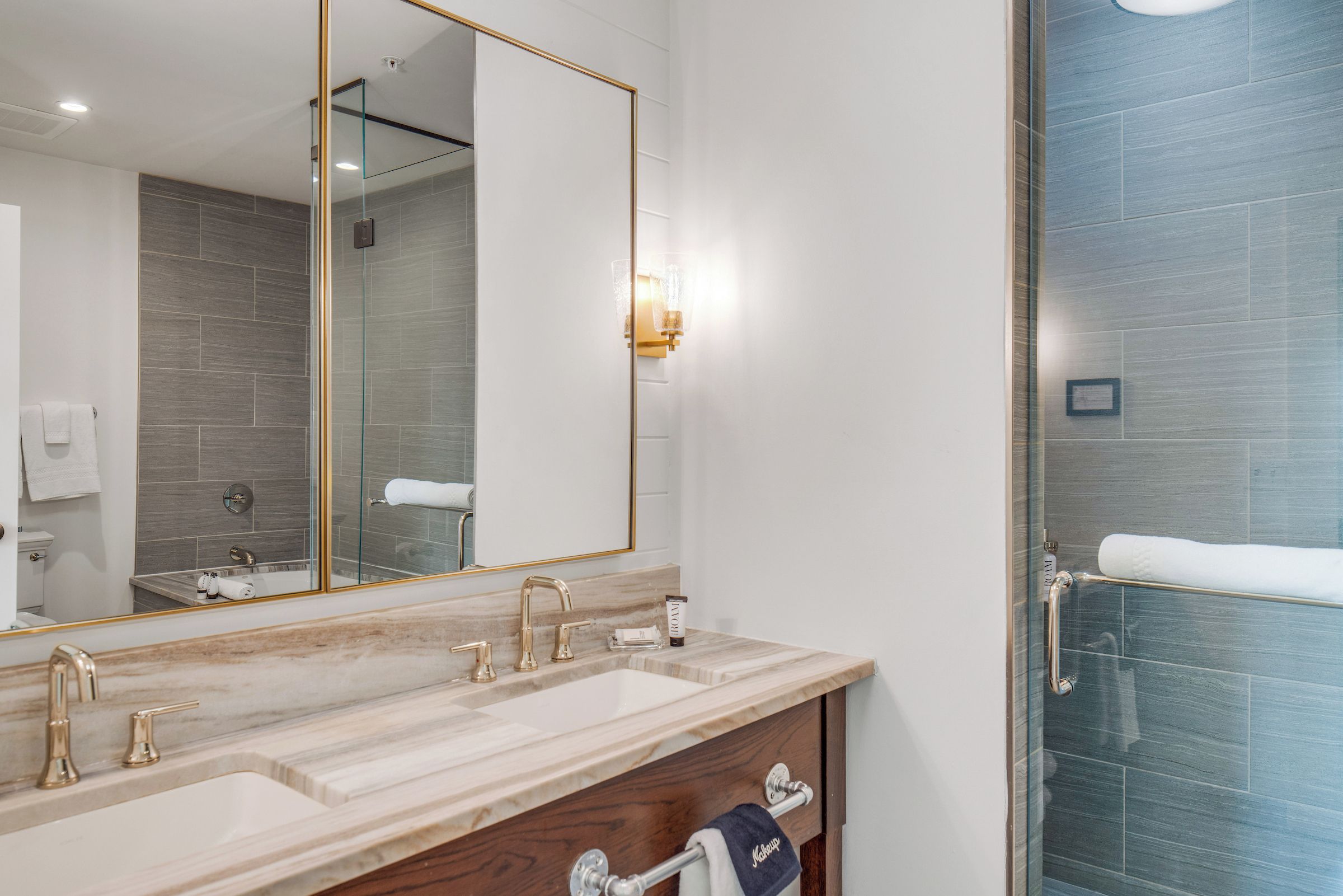 Un baño con un espejo grande, un lavamanos con dos grifos, un perchero para toallas y una zona de ducha con puerta de cristal.