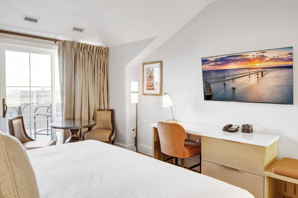 A hotel room with a bed, a desk, a chair, a table, and a view of a sunset over a bridge.