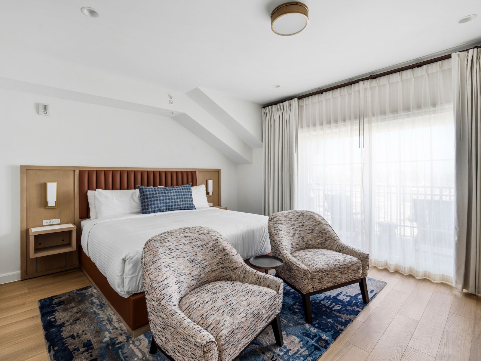A hotel room with a bed, two armchairs, a small table, and a large window with curtains.