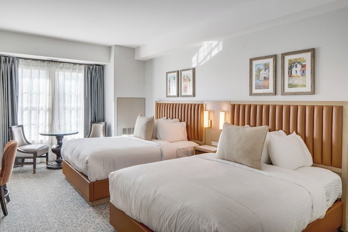 A hotel room with two beds, a sitting area with a chair and table, and artwork on the walls.