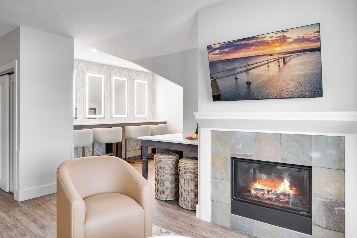 A modern living room with a fireplace, a dining area with a table and chairs, and a large painting of a sunset over a bridge.