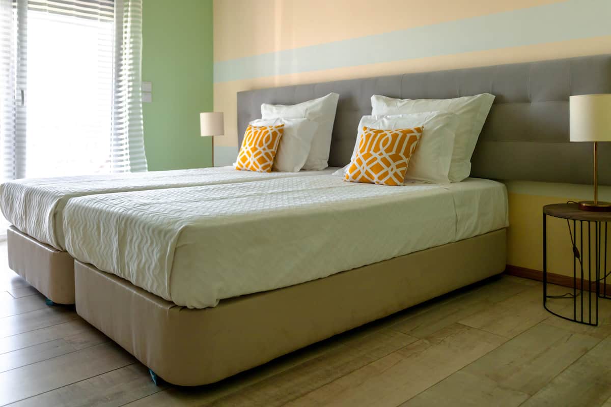 Una cama doble con almohadas blancas y anaranjadas, un cabecero tapizado en gris y una mesita de noche con una lámpara.