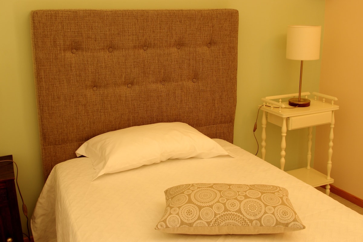 Una cama con cabecera marrón, ropa de cama blanca y una almohada estampada. Se ve una mesita de noche con una lámpara y un enchufe.