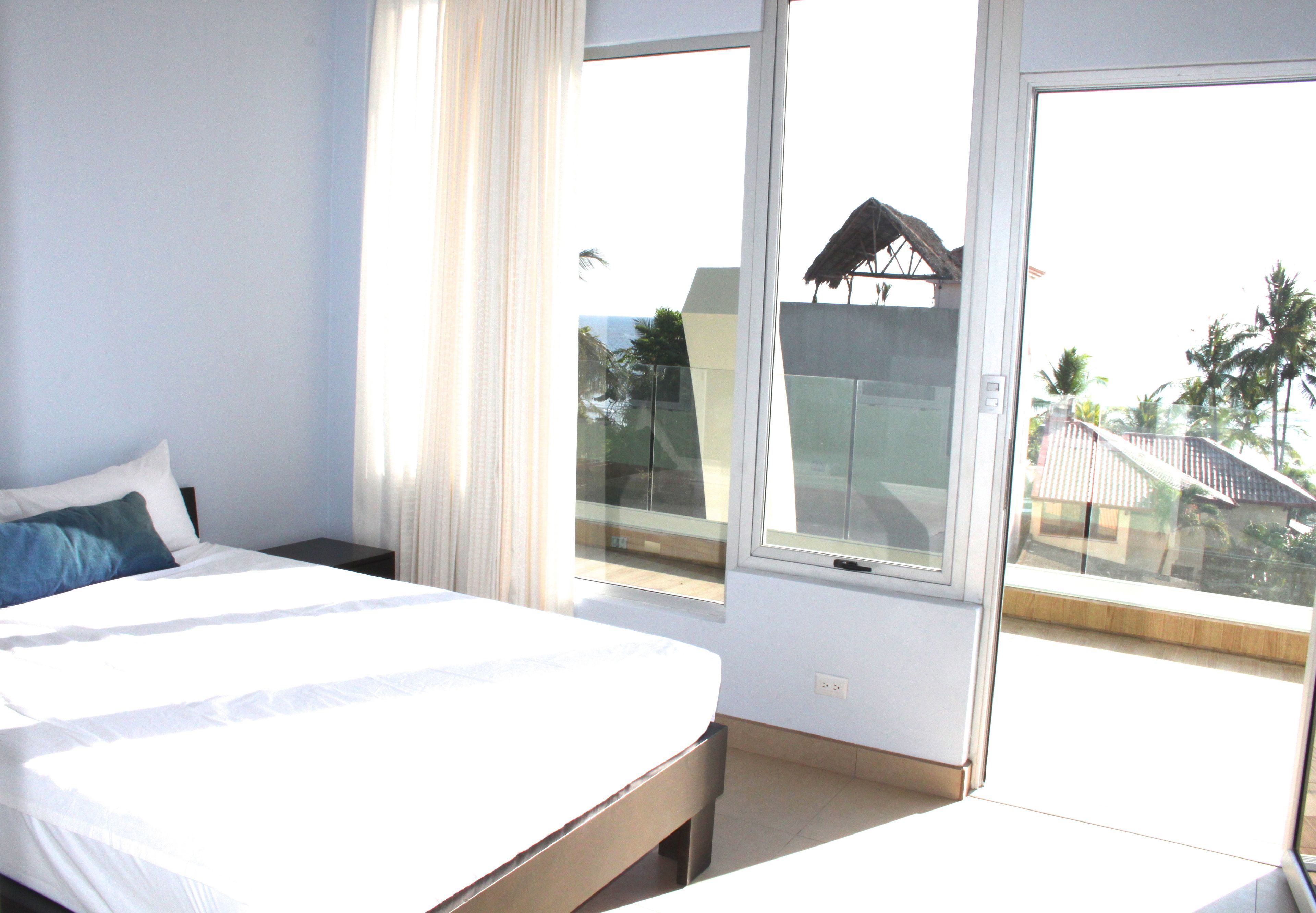 A hotel room with a bed, a nightstand, and a large window offering a view of a building and palm trees.