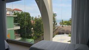 A hotel room with a large window offering a view of a coastal area, including buildings and palm trees. - Room2Board Hostel and Surf School (Jacó)