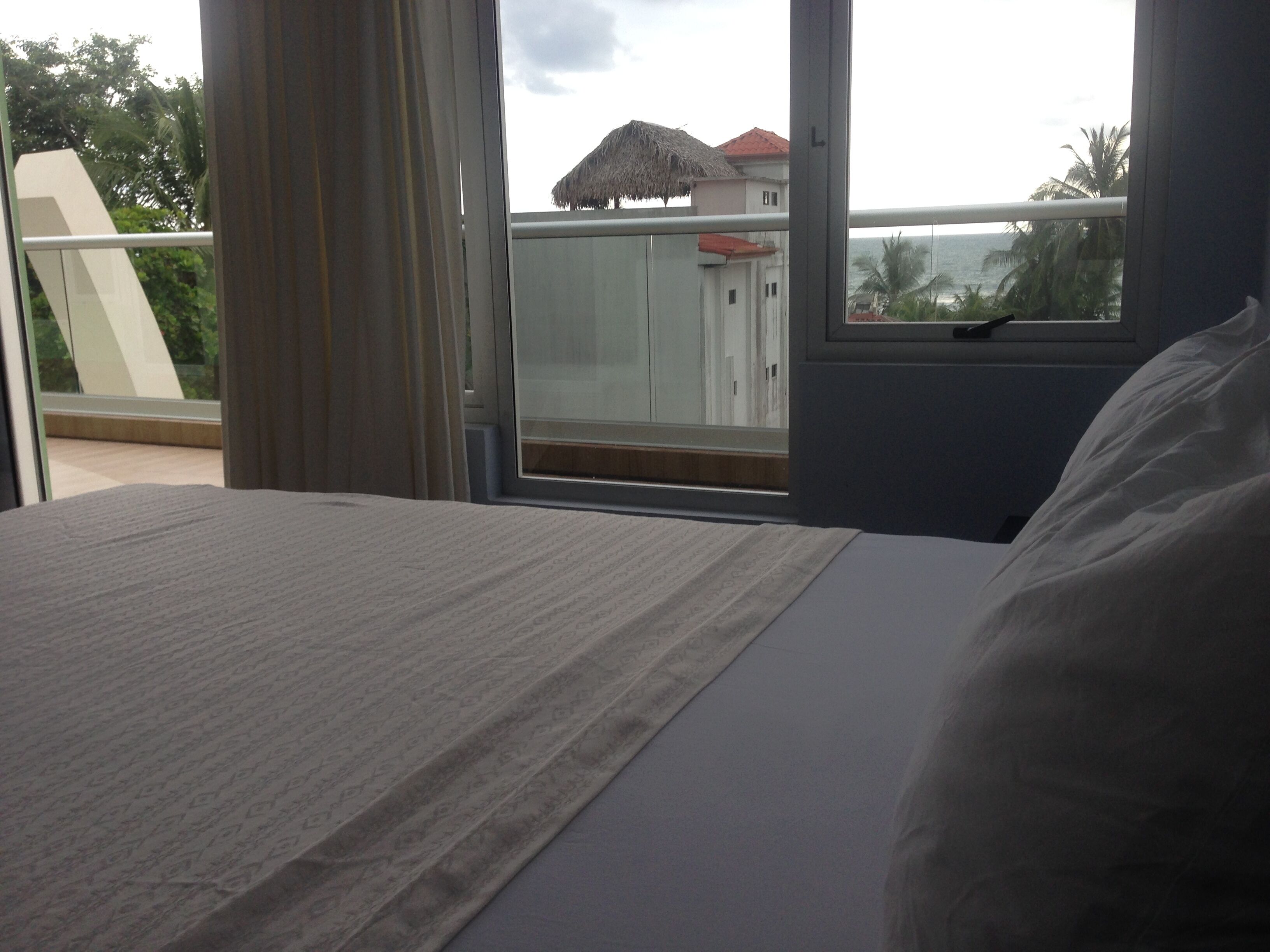 A hotel room with a bed, a window with a view of a building with a red roof, and a balcony with a thatched roof.