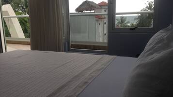 A hotel room with a bed, a window with a view of a building with a red roof, and a balcony with a thatched roof.