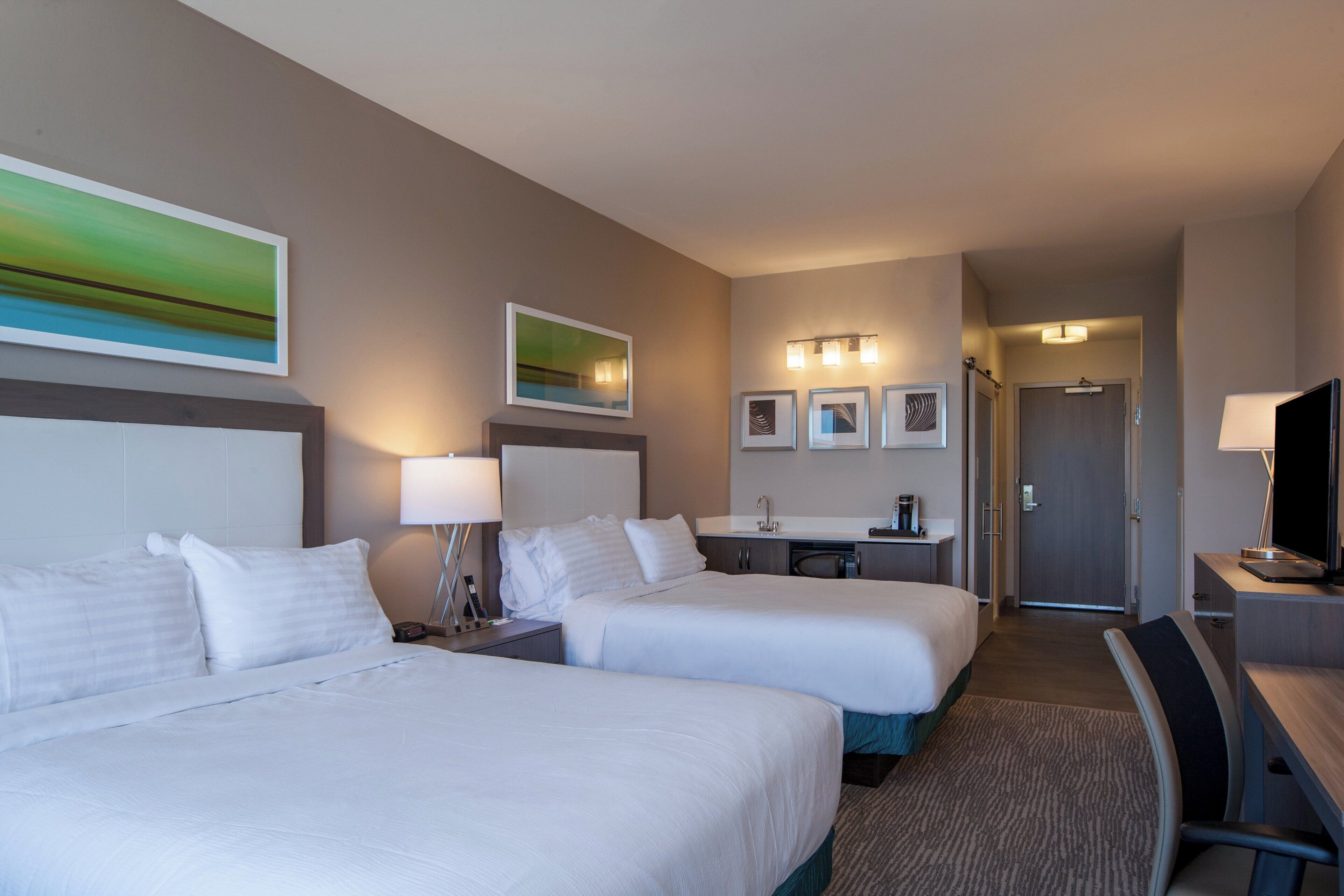 A hotel room with two beds, a desk, a chair, a TV, and a bathroom visible in the background.
