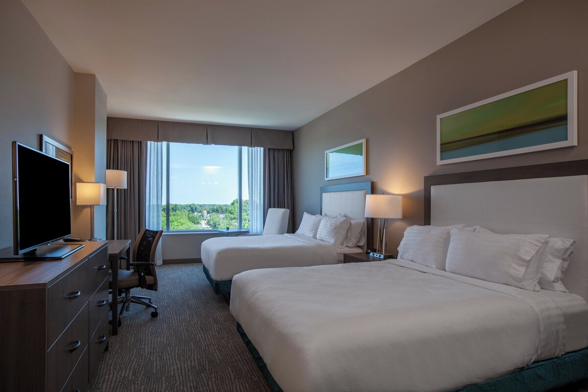 A hotel room with two beds, a desk, a television, and a view of the outdoors.