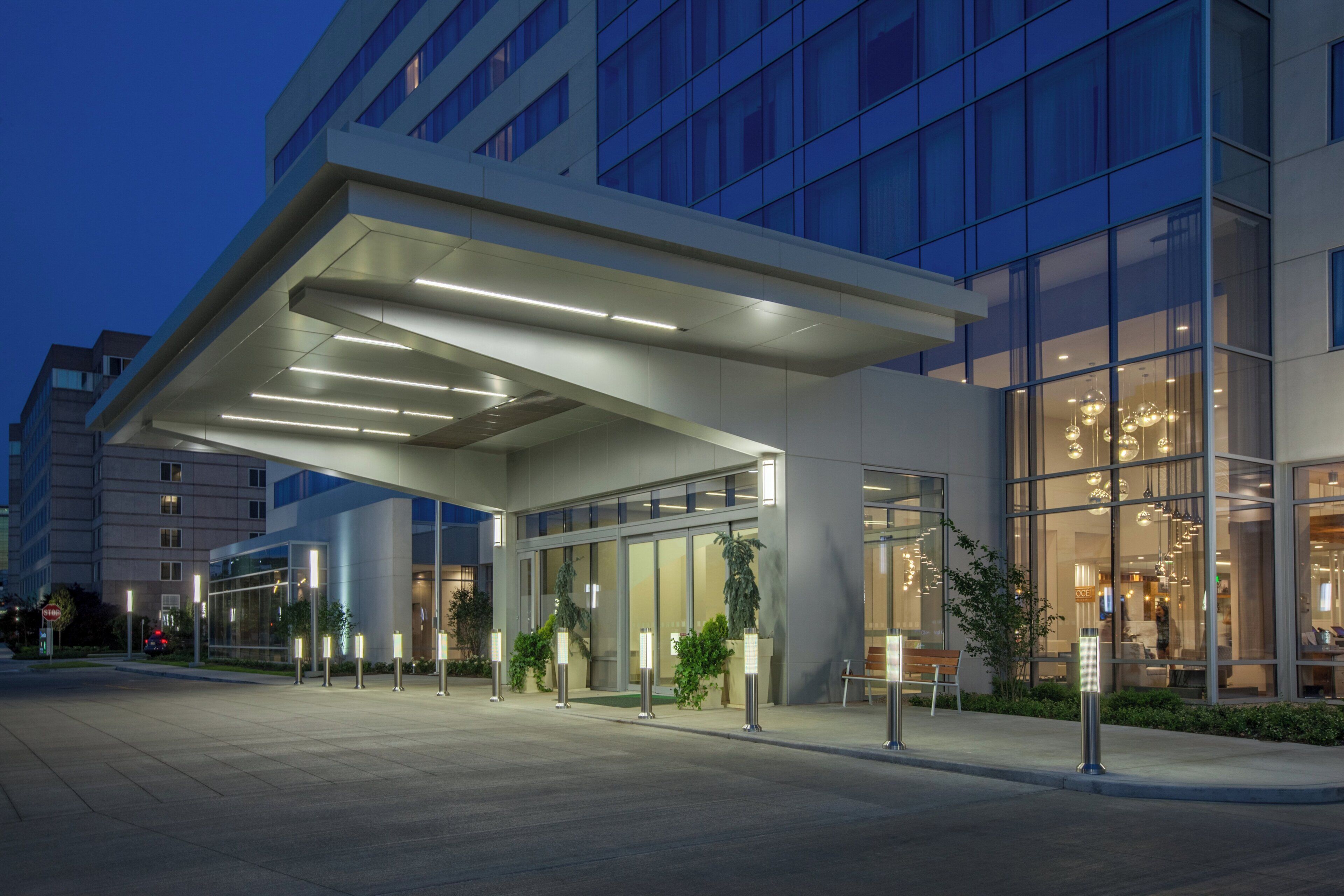 Un edificio moderno con fachada de vidrio, acceso cubierto y un área exterior bien iluminada con bancos.