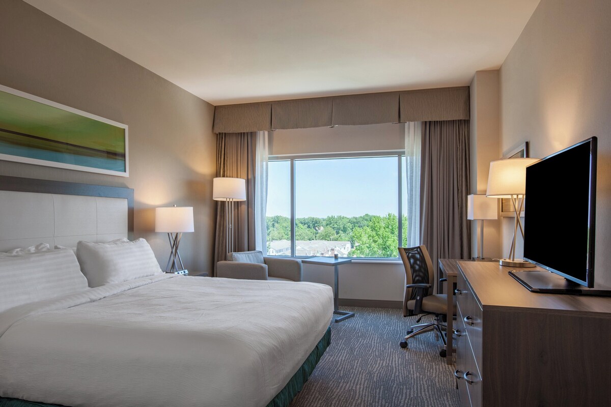 A hotel room with a large bed, a desk with a TV, a chair, and a window with curtains.