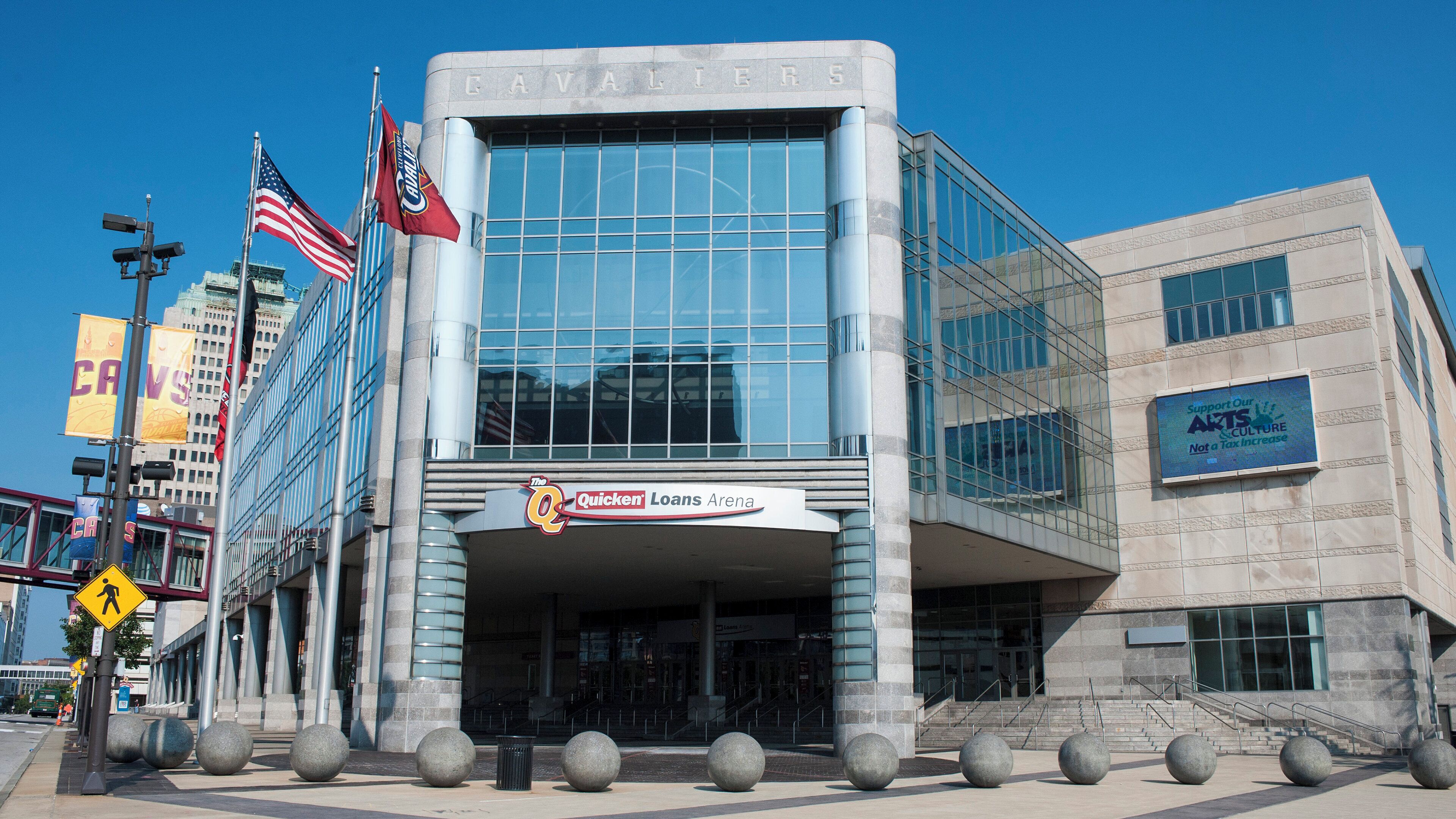 A modern building with a glass facade, a sign for 'The Q Quicken Loans Arena,' and multiple flags including the American flag.