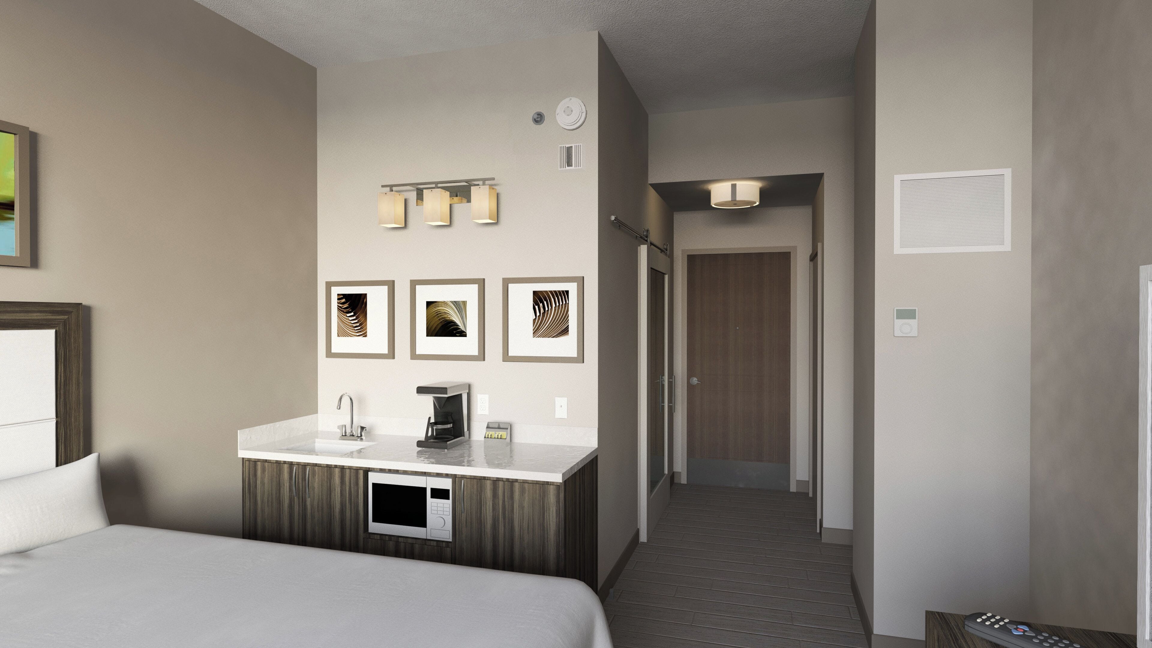 A hotel room with a bed, a minibar, a microwave, a coffee maker, a framed picture, a light fixture, a door, and a thermostat.