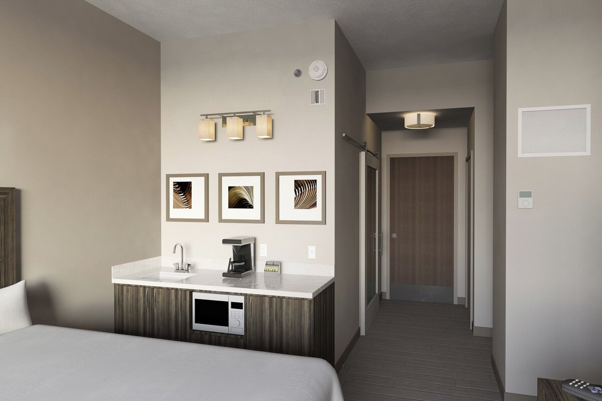 A hotel room with a bed, a minibar, a microwave, a coffee maker, a framed picture, a light fixture, a door, and a thermostat.
