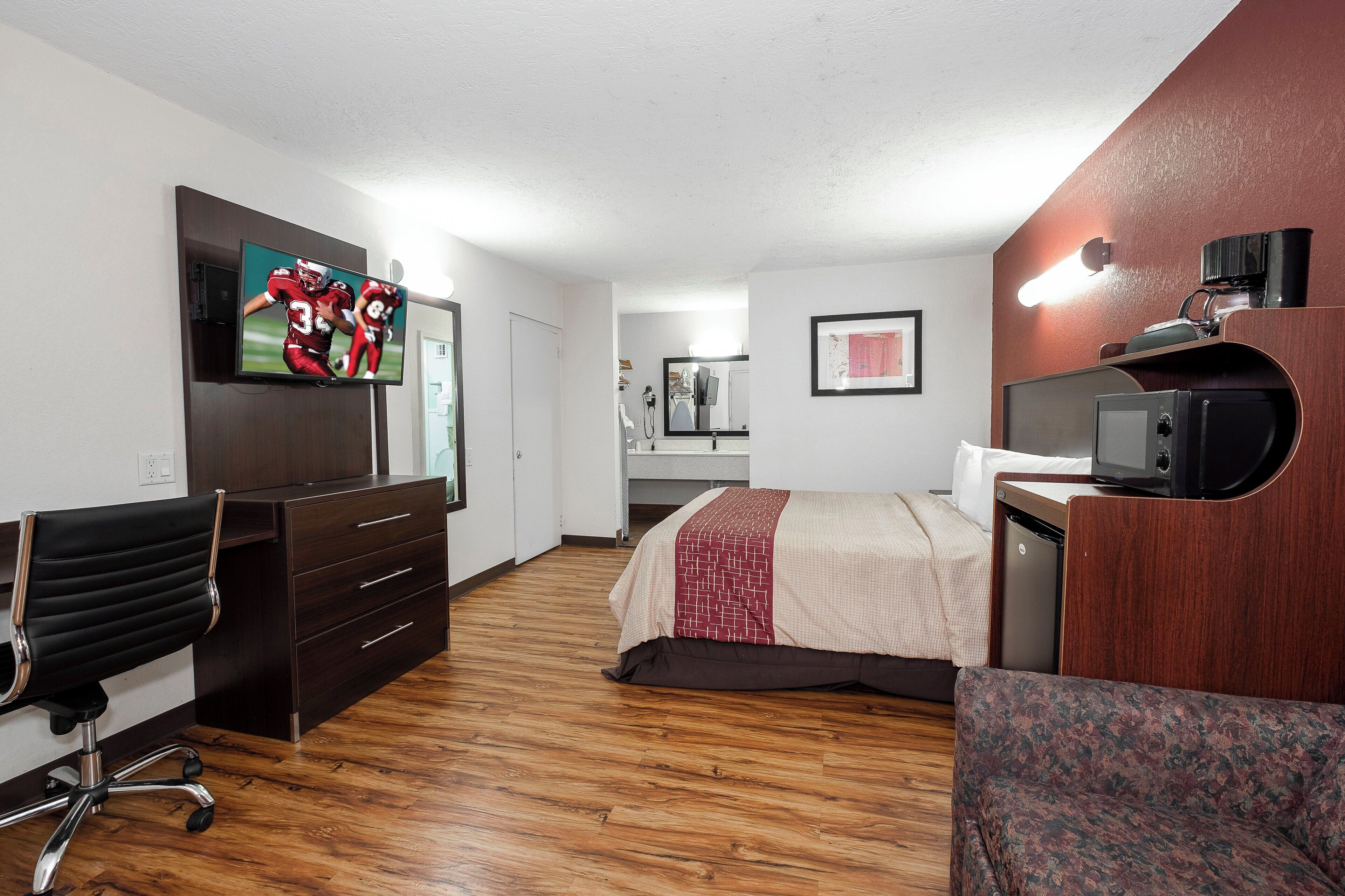 A hotel room with a wooden floor, a bed with a patterned blanket, a desk with a chair, a television, and a kitchenette area.