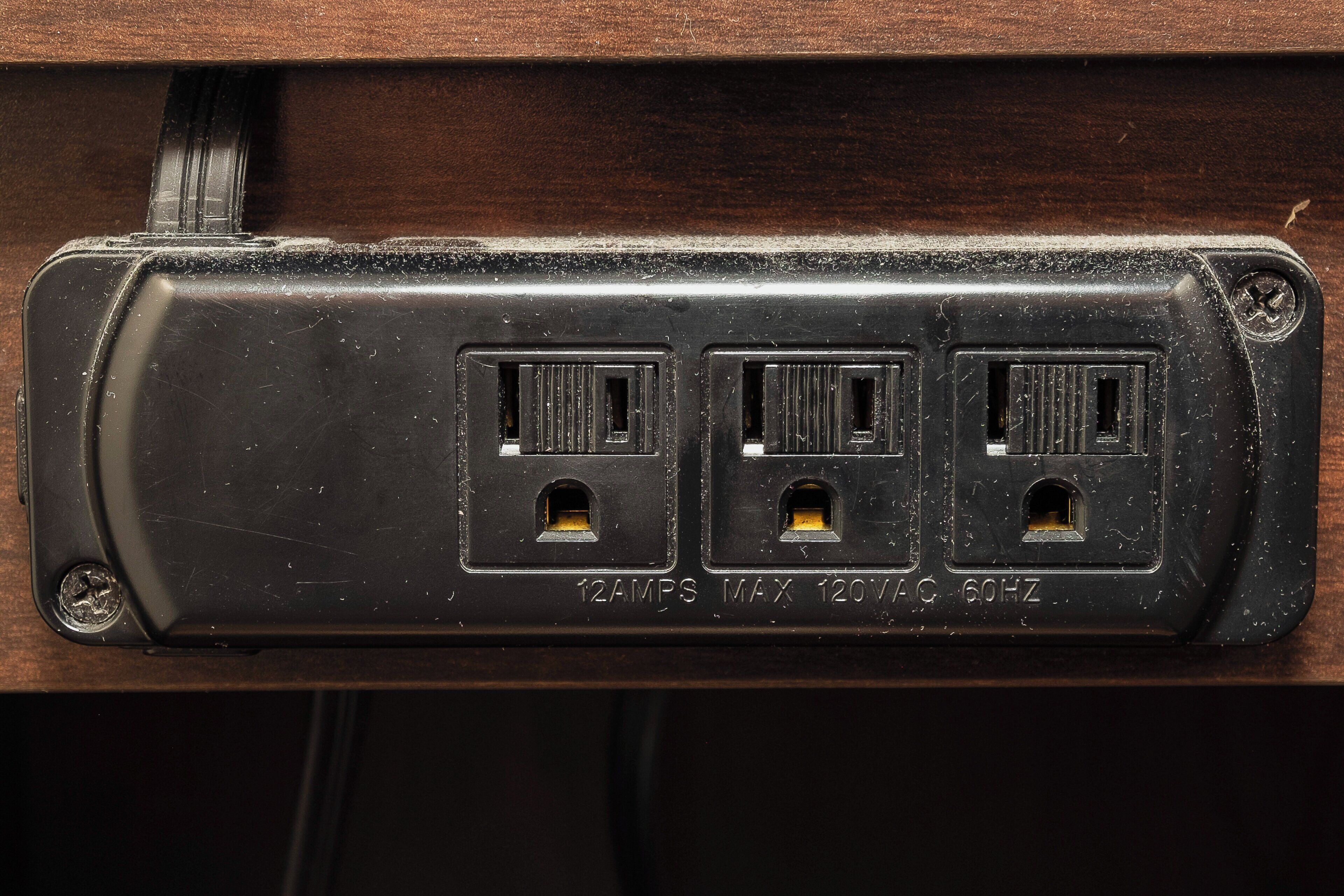 A wall-mounted electrical outlet with three sockets, a grounding screw, and a label indicating '12AMP 120VAC 60HZ'.