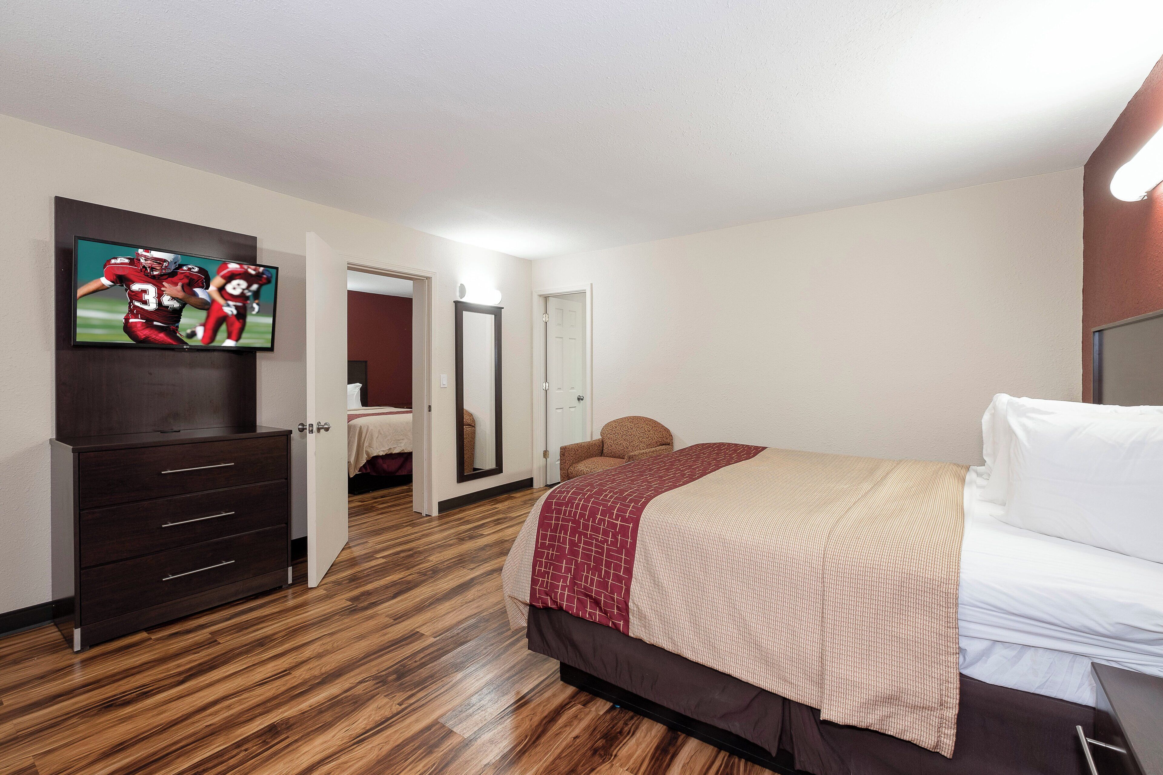 A hotel room with a bed, a TV mounted on the wall, a wooden floor, and a door leading to another room.