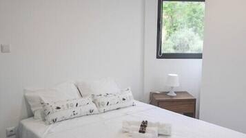 A neatly made bed with white linens and patterned pillows, a bedside table with a lamp, and a window with a view of greenery.