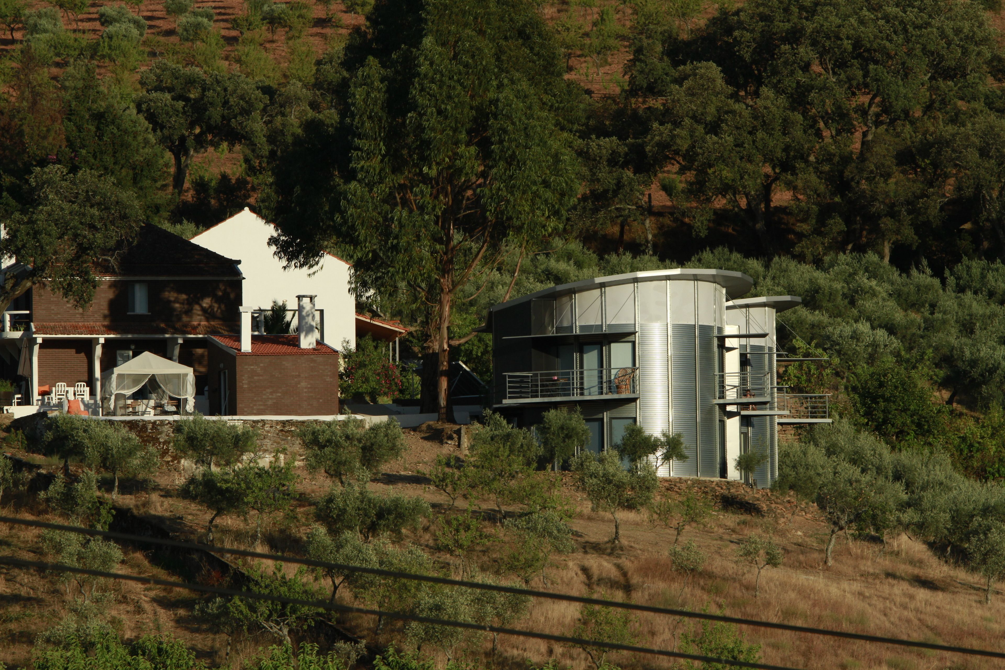 Una casa moderna con un gran árbol en primer plano, una casa más pequeña con fachada blanca y una estructura curva con balcones.