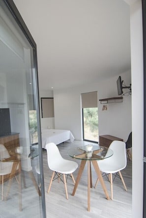 Une chambre moderne avec une cloison en verre, un lit, une petite table avec des chaises et un téléviseur fixé au mur.