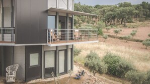 Ein modernes, zweistöckiges Haus mit Balkon, großen Fenstern und Blick auf eine trockene, grasbewachsene Landschaft mit Bäumen.