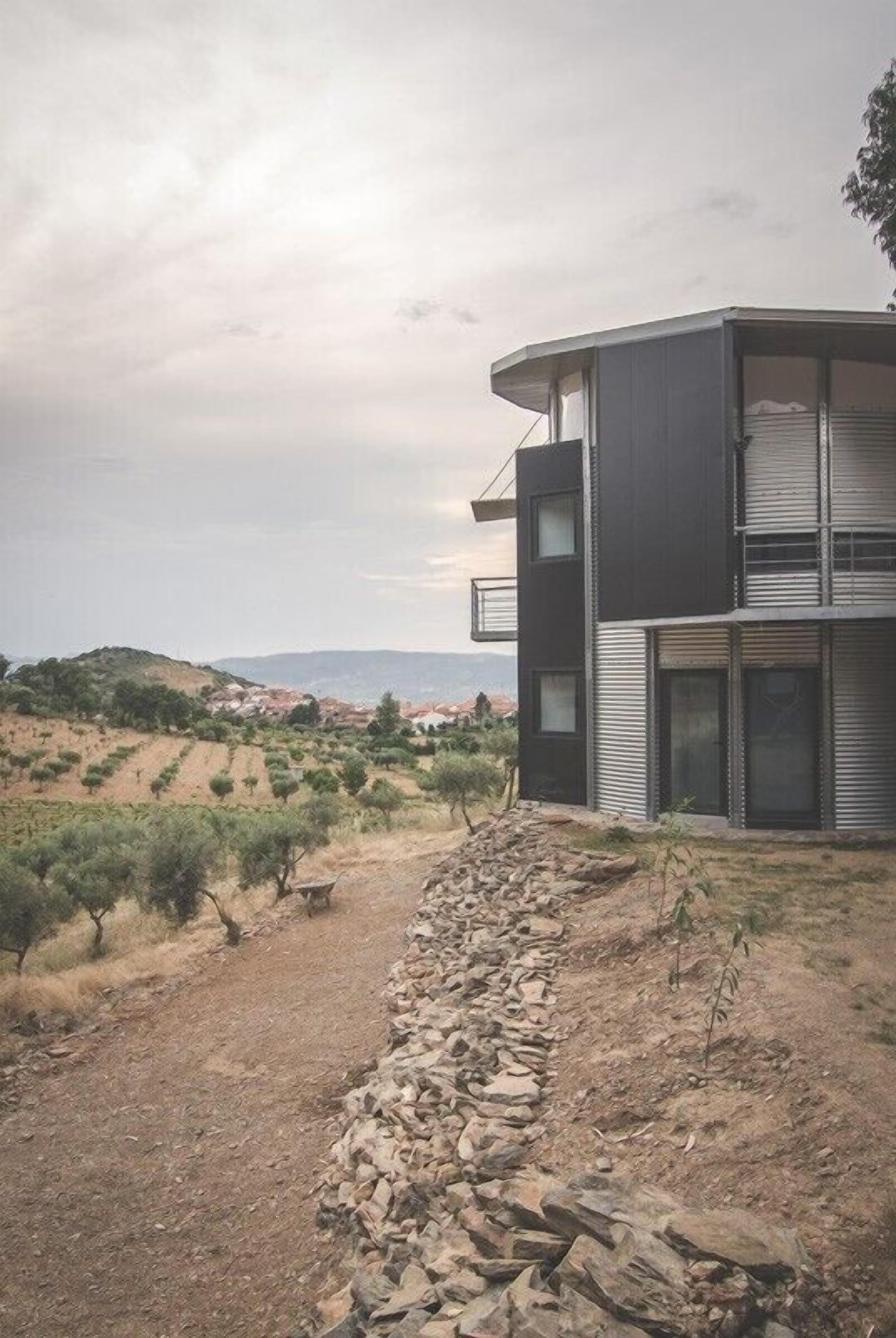 Um edifício moderno de dois andares, com um caminho de pedra que o leva até ele, rodeado por uma paisagem acidentada com vegetação escassa.