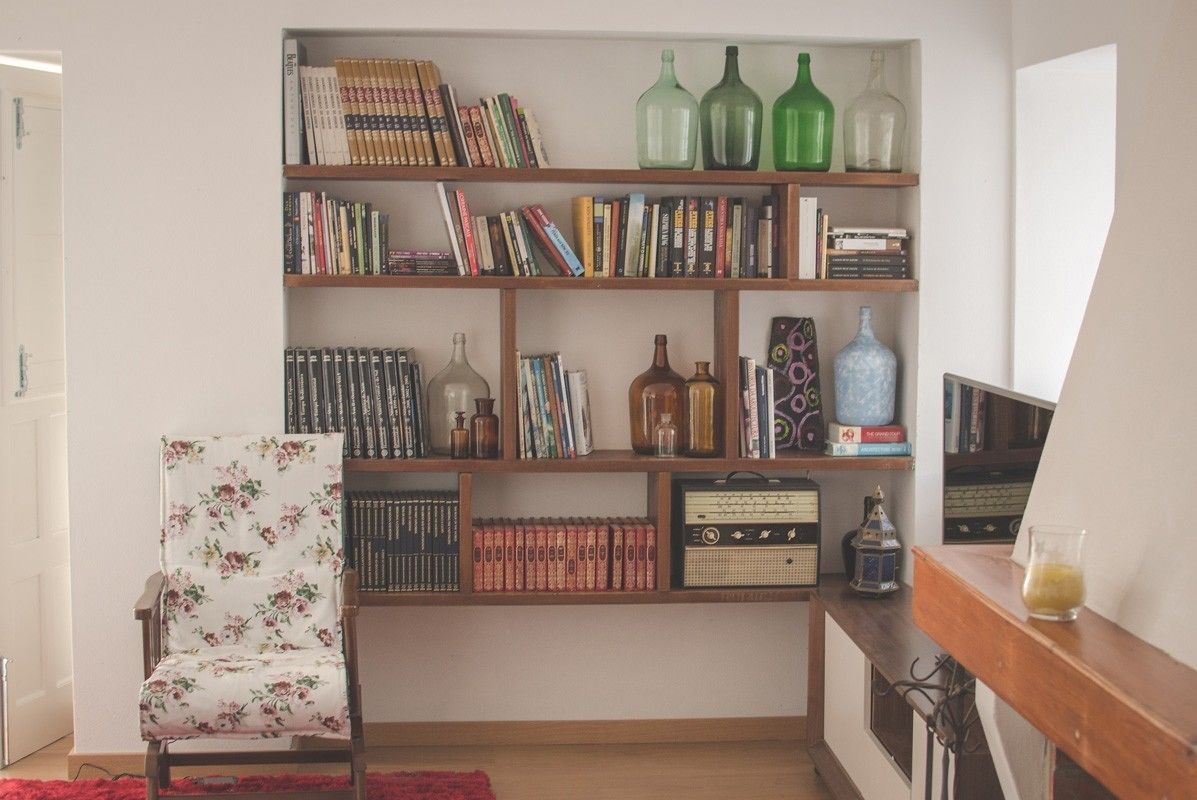 Una habitación con una estantería de madera llena de libros y objetos decorativos, un sillón floral y una alfombra roja.