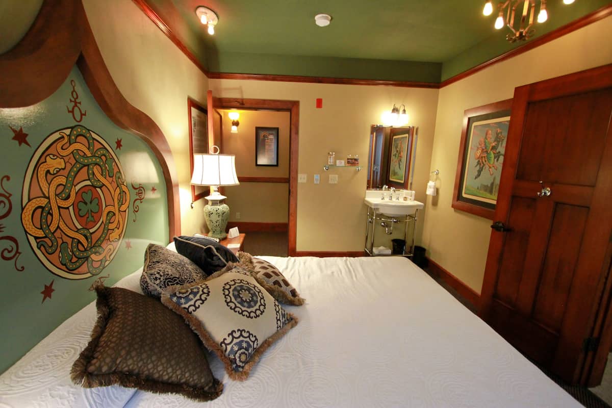 A hotel room with a large bed, a bedside table with a lamp, a bathroom with a sink and mirror, and a door with a wooden frame.