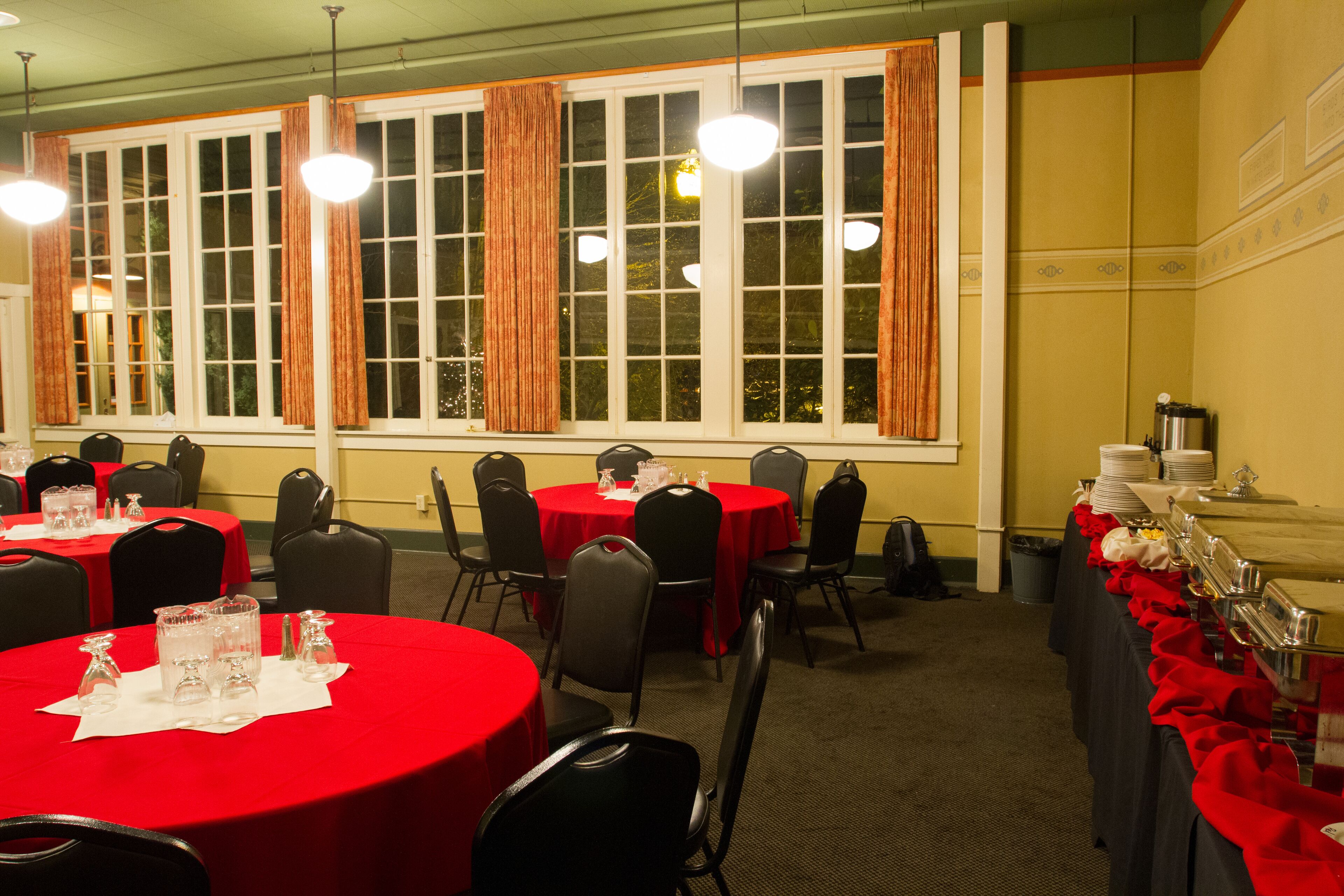 Une salle de banquet avec des tables rondes recouvertes de nappes rouges, des chaises noires et de grandes fenêtres ornées de rideaux.