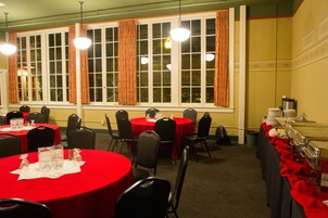 Une salle de banquet avec des tables rondes recouvertes de nappes rouges, des chaises noires et de grandes fenêtres ornées de rideaux.