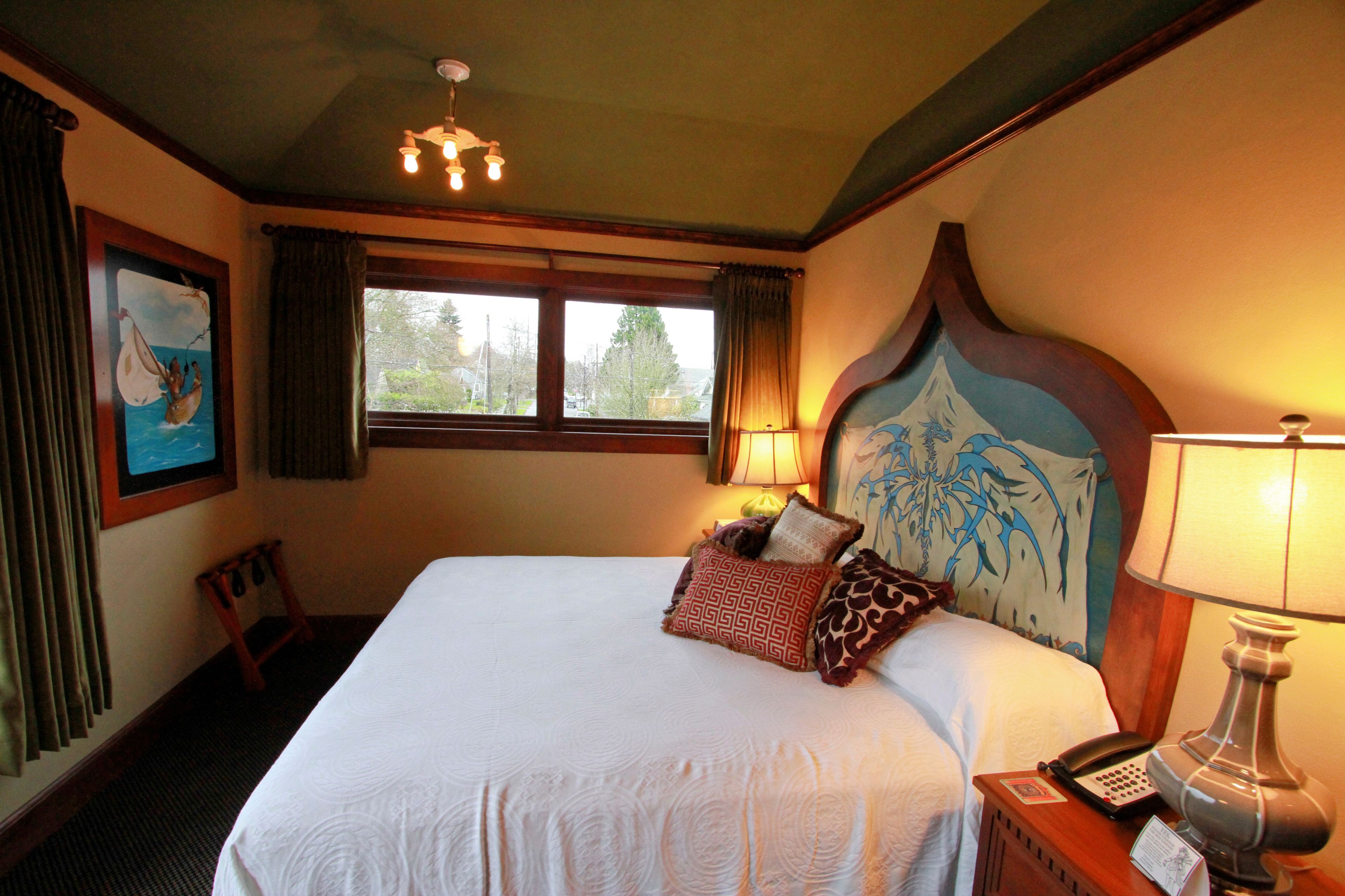 Una habitación de hotel con una cama grande, cabecera de madera, ventana con cortinas, mesita de noche con lámpara y un cuadro en la pared.