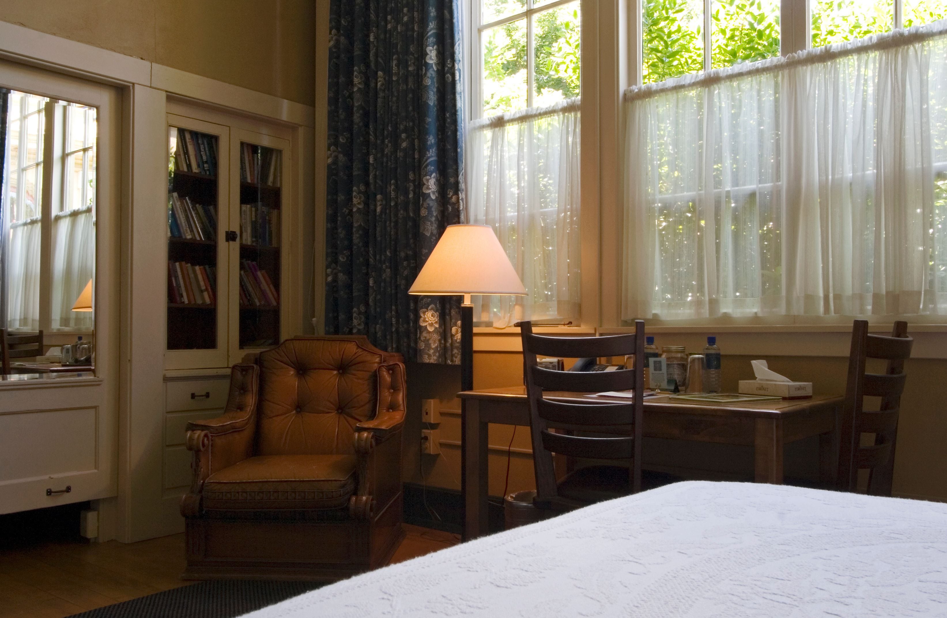 A hotel room with a bed, a desk with a lamp, a chair, a bookshelf, and a window with curtains.