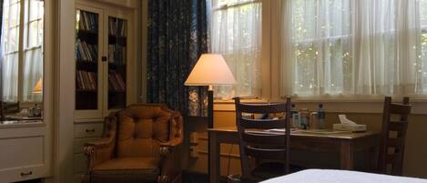 A hotel room with a bed, a desk with a lamp, a chair, a bookshelf, and a window with curtains.