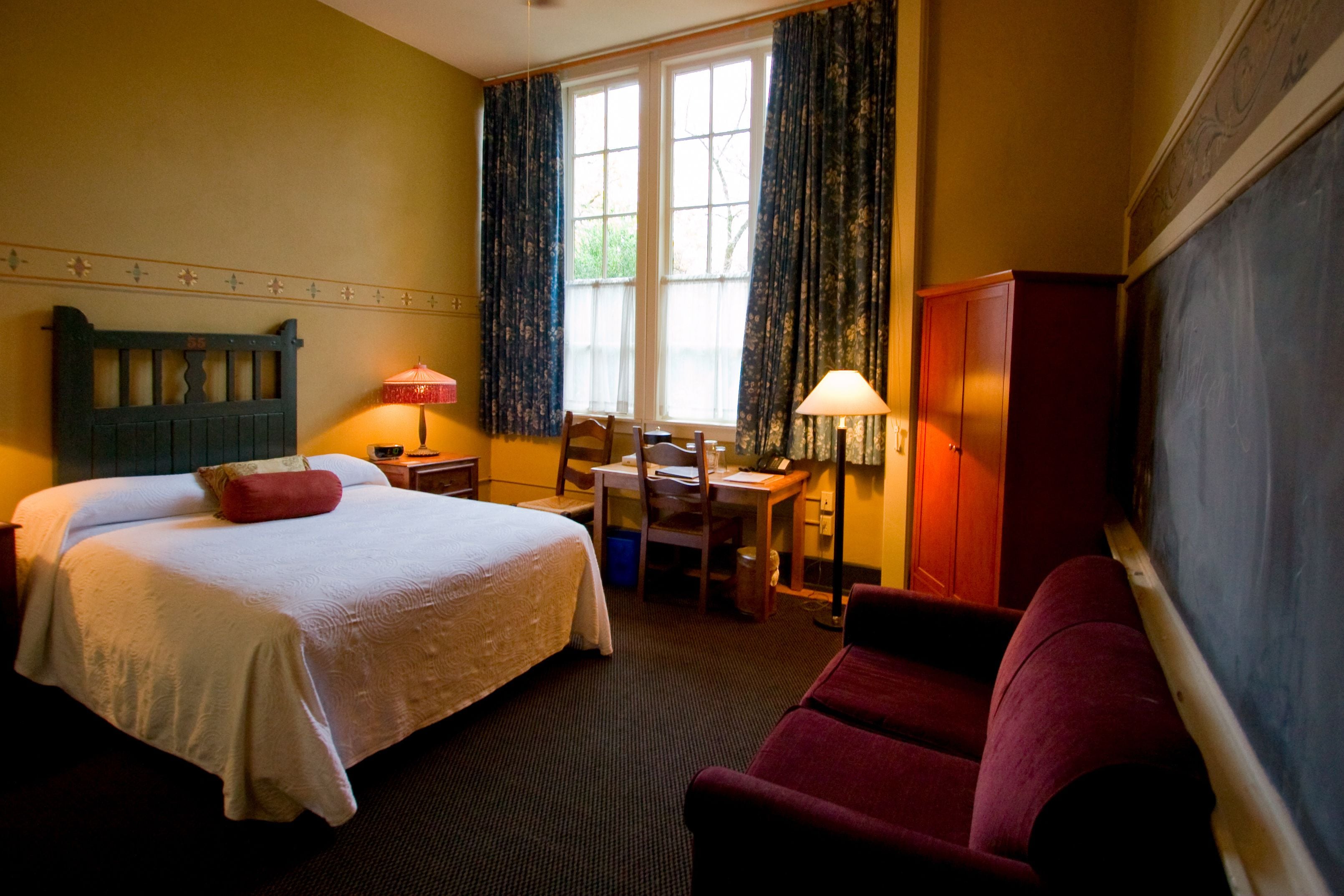 A hotel room with a bed, a desk, a chair, a red armchair, and a wardrobe.