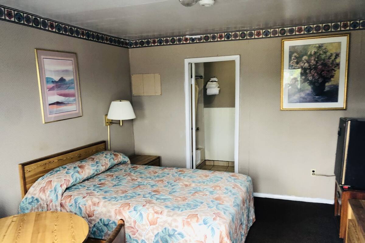 Una habitación de hotel con una cama, una mesita de noche, una silla, una lámpara, un televisor y una puerta que da a un baño.