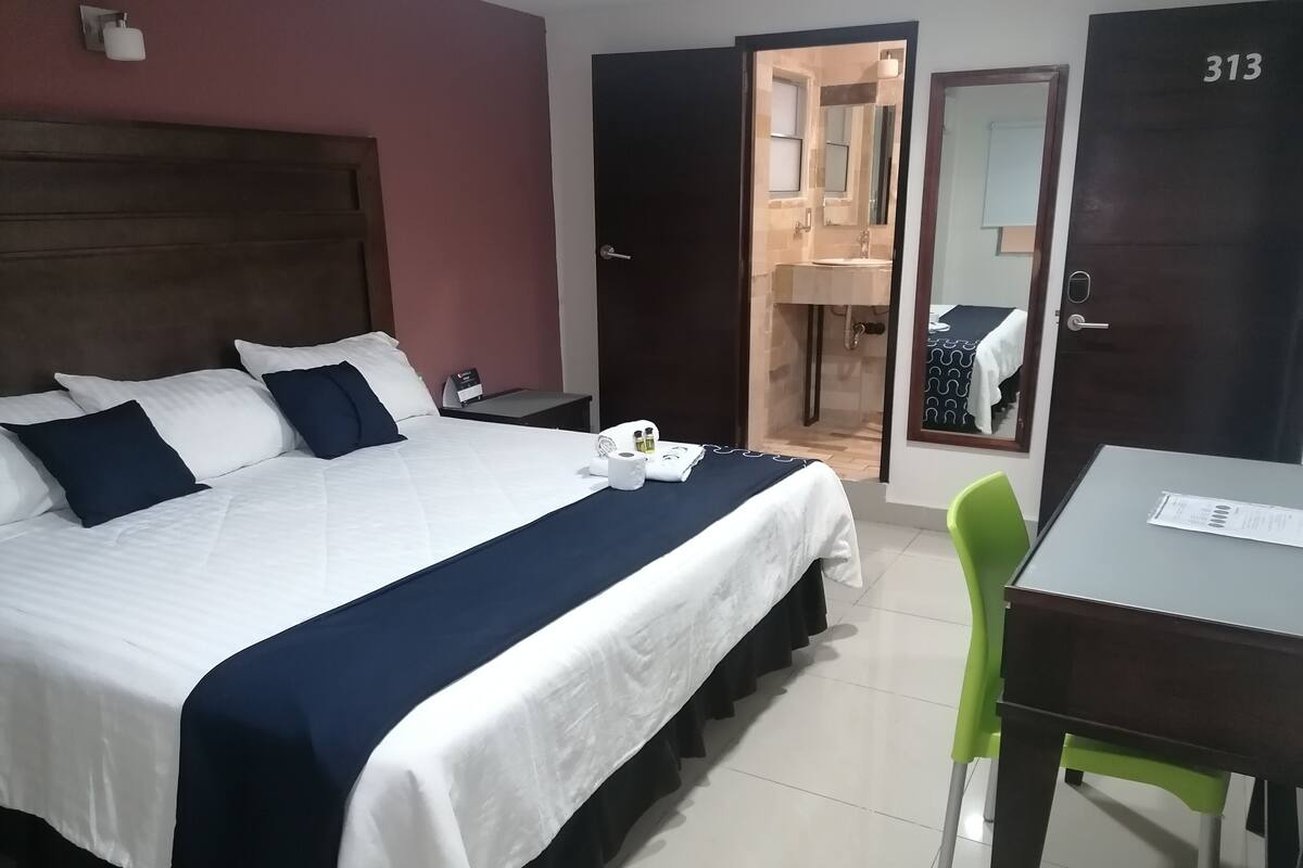 A hotel room with a bed, a desk, a chair, and a bathroom visible through an open door.