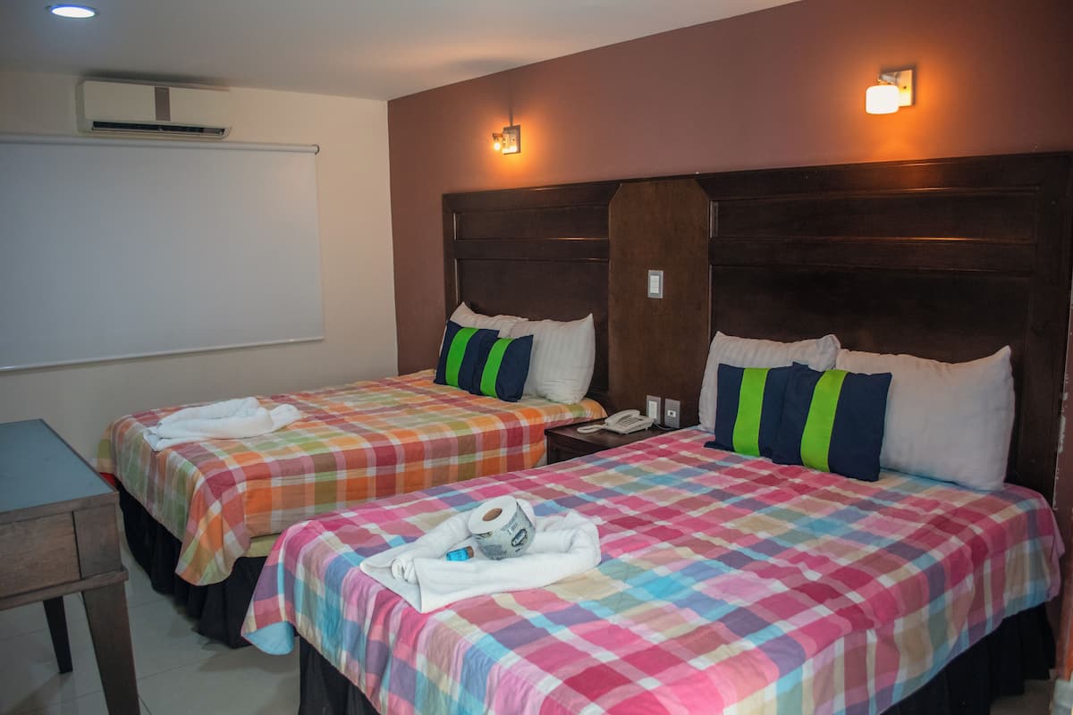 A hotel room with two beds, each with colorful bedding and pillows, a bedside table with a phone, and a wall-mounted air conditioner.