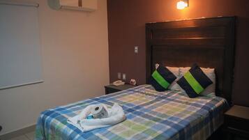 A hotel room with a checkered bedspread, a bedside table with a lamp, and a wall-mounted air conditioner.