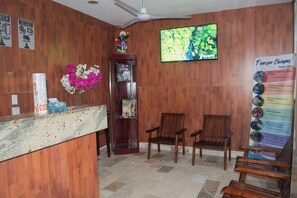 A reception area with a wooden counter, two wooden chairs, a ceiling fan, a TV screen, and a poster with tour information. - Granda Inn (Tuxtla Gutierrez)