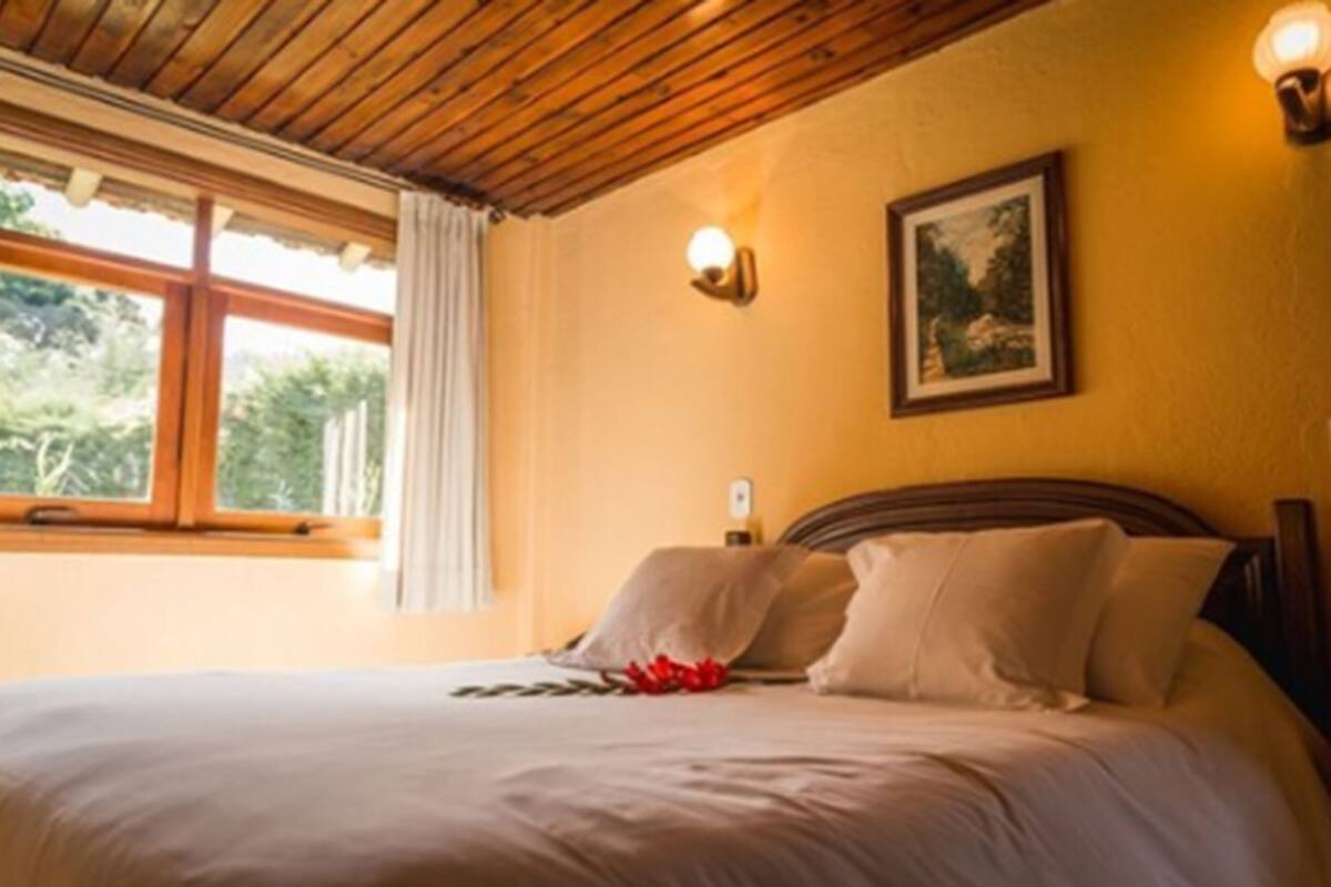 Un dormitorio con techo de madera, una cama con ropa de cama blanca, una mesita de noche, una lámpara de pared y un cuadro enmarcado.