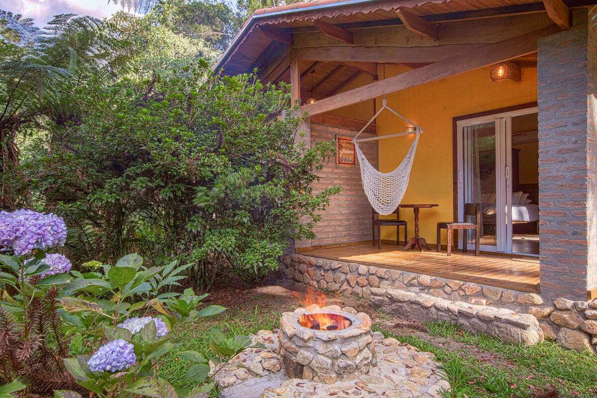 Un rincón exterior acogedor con un hogar de piedra, una hamaca y vista a un dormitorio a través de un ventanal.
