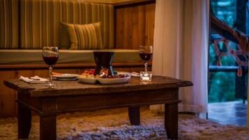 Una mesa de madera con un plato de fruta, dos copas de vino tinto y una cafetera en una habitación acogedora con un banco tapizado de color verde y vistas a un bosque a través de una ventana.