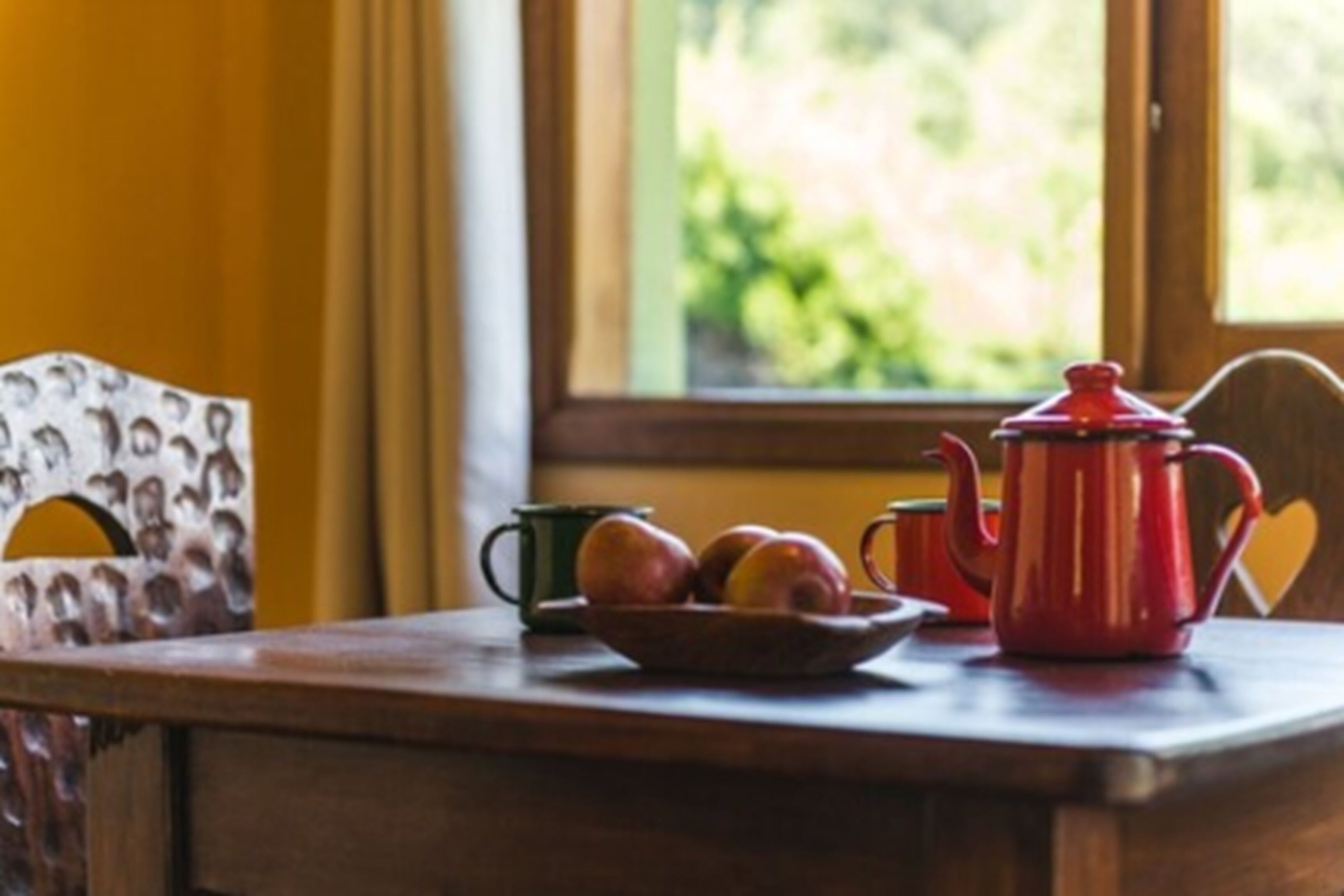 A wooden table with a bowl of fruit, a green mug, and a red teapot.