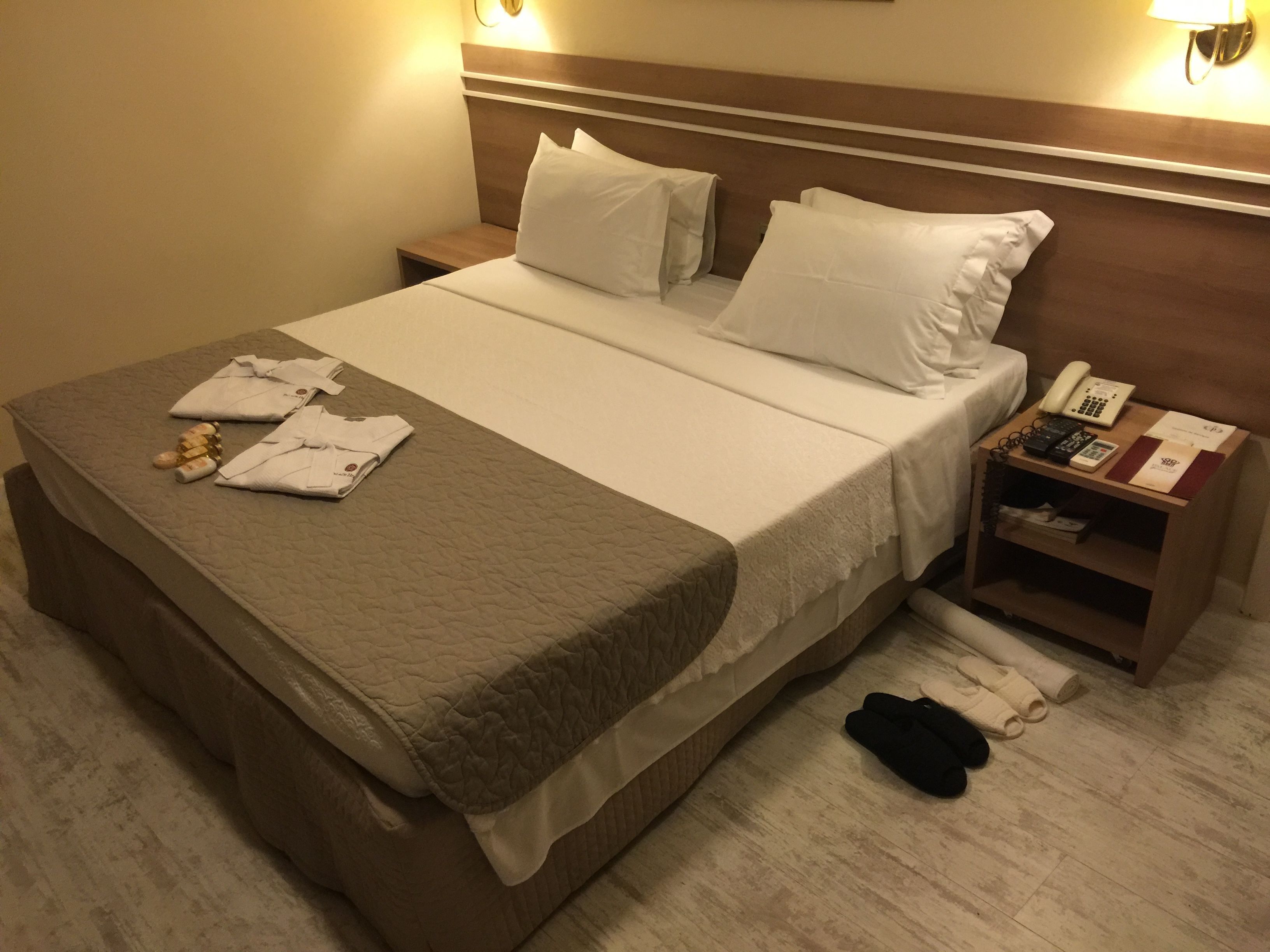 A neatly made bed with white linens, a bedside table with a phone and a book, and a pair of slippers on the floor.